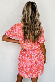 Donna con abito estivo floreale rosa e arancione, capelli ondulati biondi, sfondo bianco. Moda femminile, abbigliamento casual elegante.