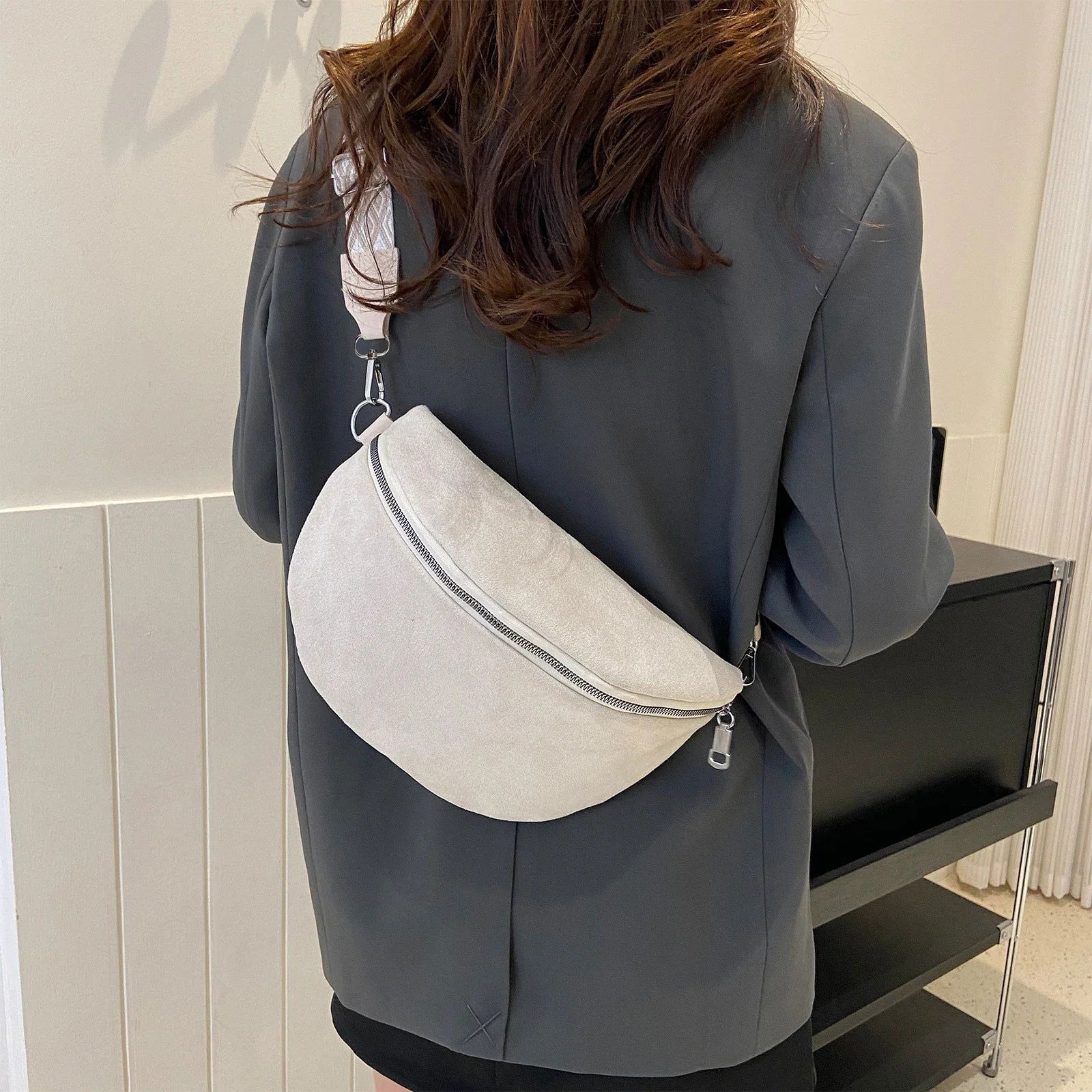Donna con giacca grigia indossa marsupio bianco in pelle scamosciata, chiusura zip, tracolla regolabile. Accessorio moda casual elegante.