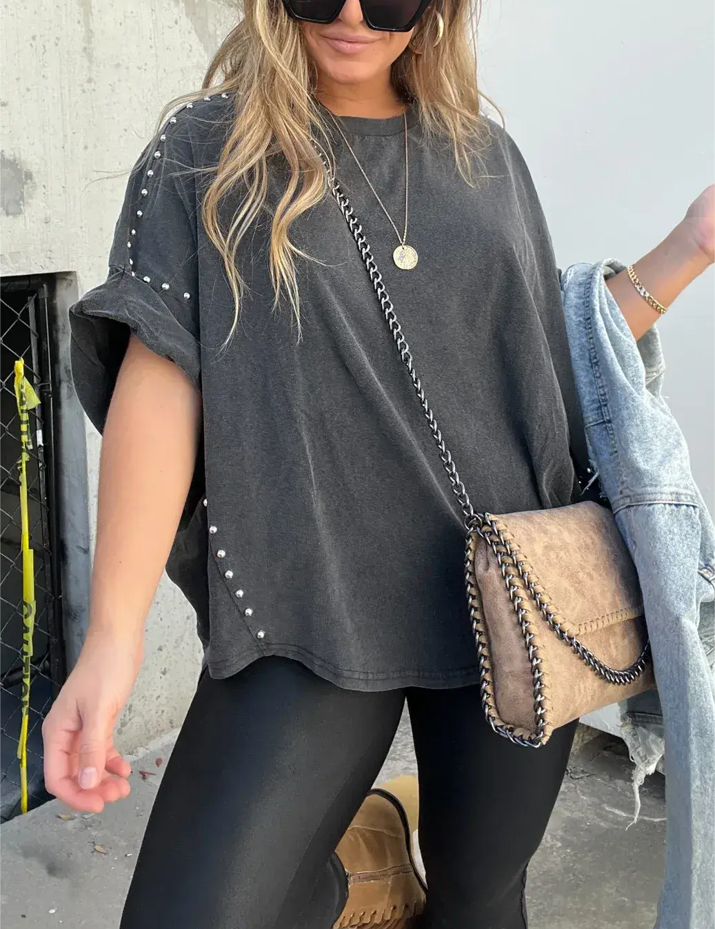 Donna con maglietta oversize nera con borchie, leggings neri, borsa a tracolla beige e occhiali da sole. Moda casual urbana.