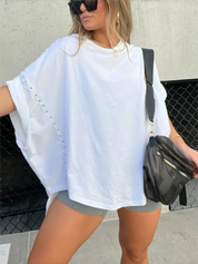 Donna con maglietta bianca oversize con borchie, pantaloncini grigi, borsa a tracolla nera e occhiali da sole. Moda casual estiva urbana.