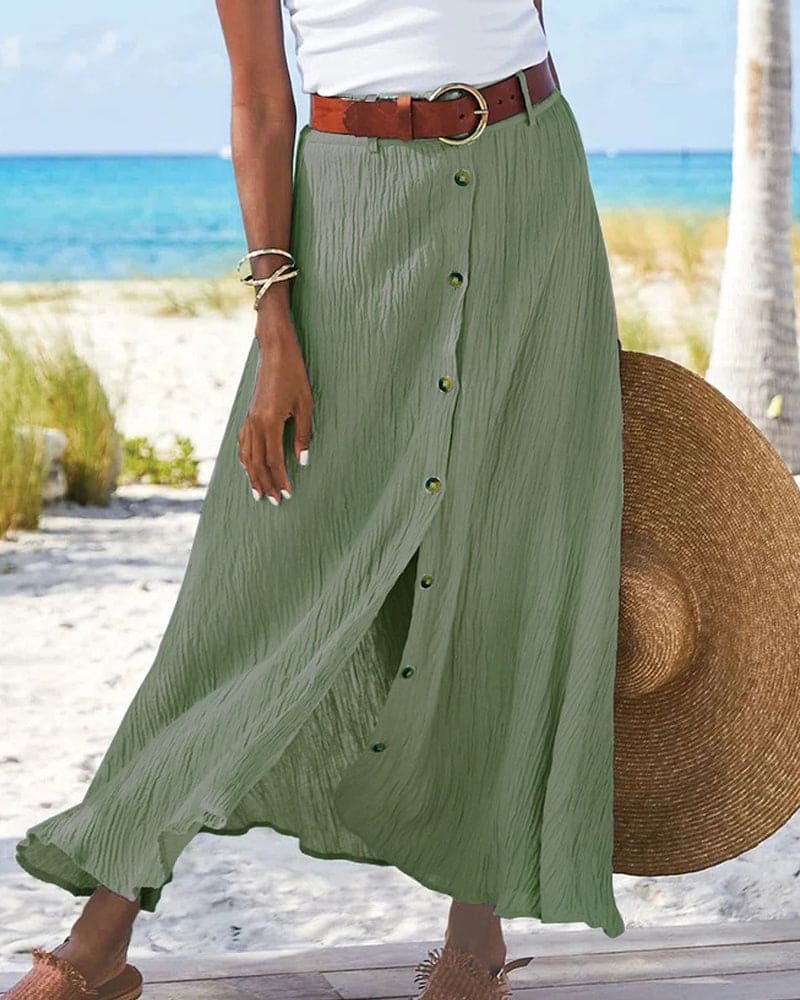 Gonna lunga verde con bottoni, cintura marrone, indossata su spiaggia estiva. Moda donna, abbigliamento estivo, look casual elegante.