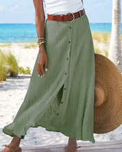 Gonna lunga verde con bottoni, cintura marrone, indossata su spiaggia estiva. Moda donna, abbigliamento estivo, look casual elegante.