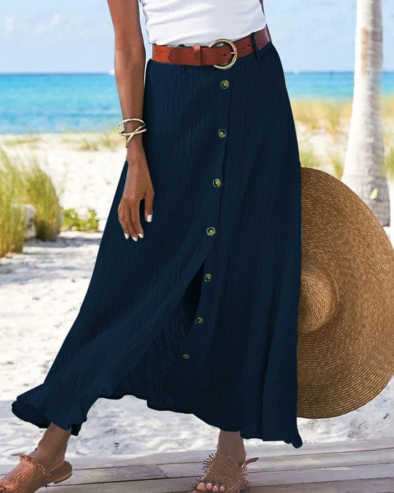 Gonna lunga blu con bottoni, cintura marrone, indossata su spiaggia estiva. Moda donna, abbigliamento estivo, stile casual elegante.