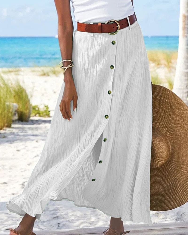 Gonna lunga bianca con bottoni verdi, cintura marrone, indossata su spiaggia tropicale. Moda estiva donna, abbigliamento casual elegante.