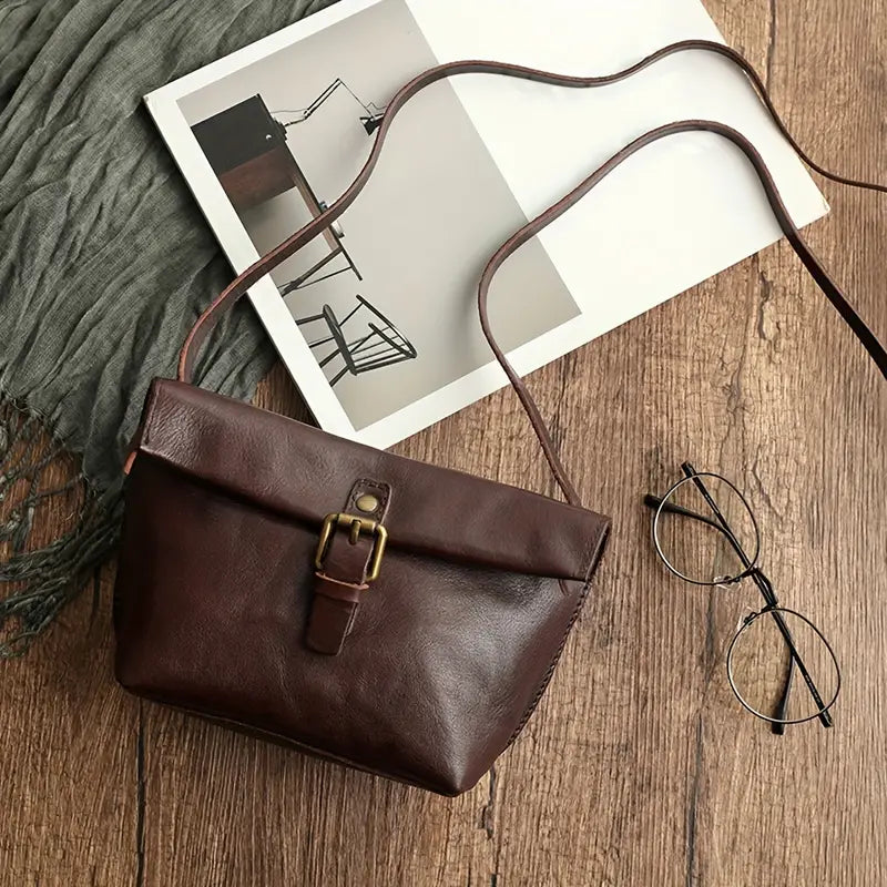 Borsa a tracolla in pelle marrone su tavolo di legno, accanto a occhiali e rivista. Elegante accessorio moda donna, stile vintage.