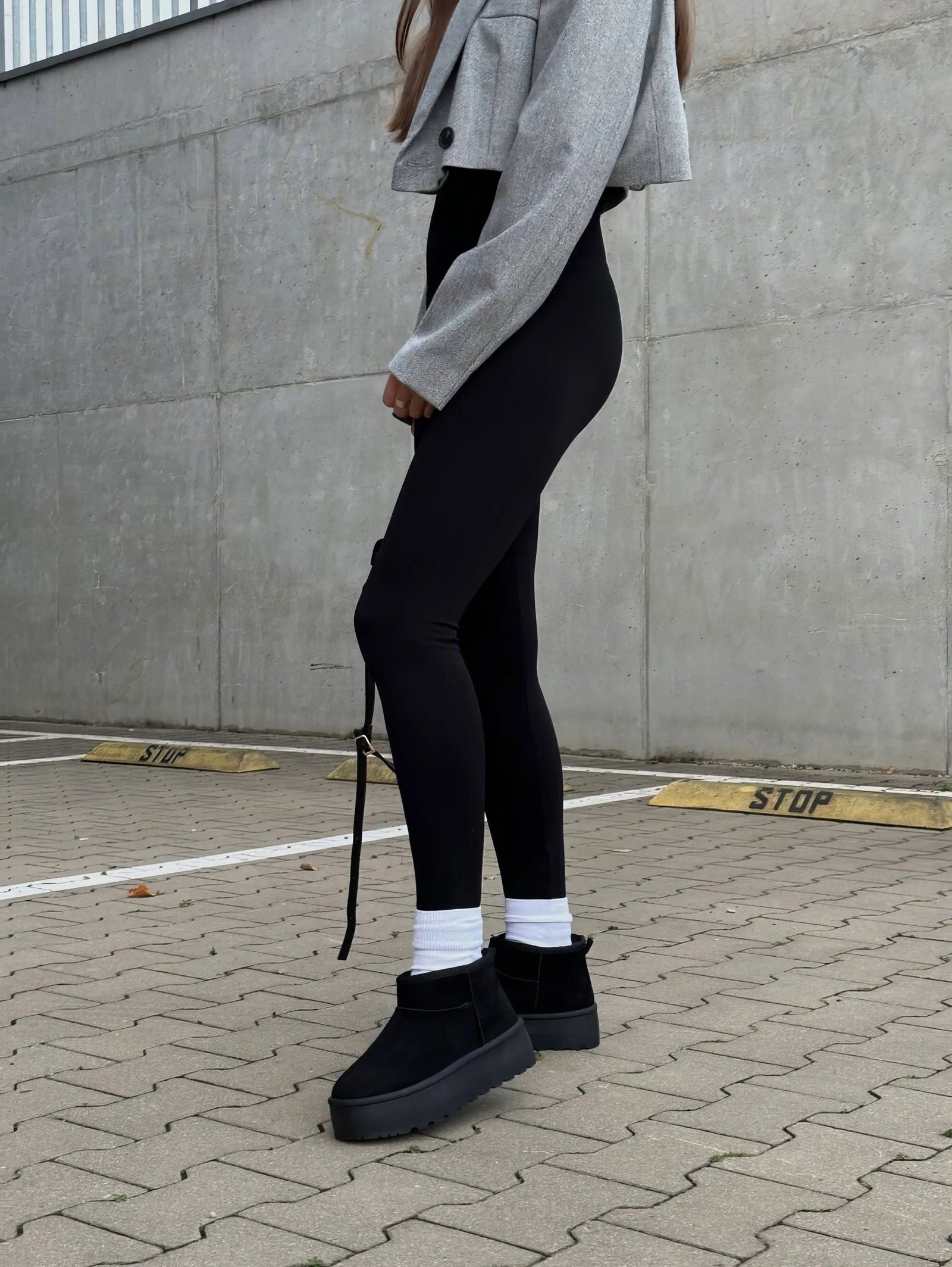 Donna con leggings neri e stivali invernali neri, giacca corta grigia, su sfondo urbano. Moda casual autunnale, abbigliamento comodo e trendy.