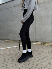 Donna con leggings neri e stivali invernali neri, giacca corta grigia, su sfondo urbano. Moda casual autunnale, abbigliamento comodo e trendy.
