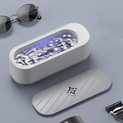 Dispositivo di pulizia a ultrasuoni bianco con coperchio, occhiali da sole e rasoio su sfondo grigio. Tecnologia avanzata per igienizzazione efficace.