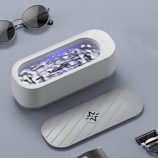 Dispositivo di pulizia a ultrasuoni bianco con coperchio, occhiali da sole e rasoio su sfondo grigio. Tecnologia avanzata per igienizzazione efficace.