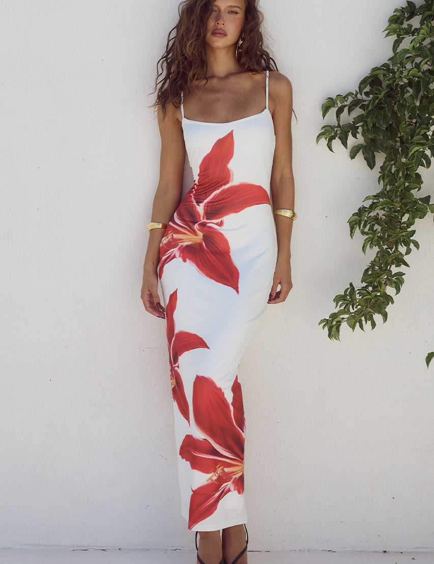 Abito lungo estivo bianco con stampa floreale rossa, spalline sottili, indossato da donna su sfondo bianco con pianta verde. Moda femminile elegante.