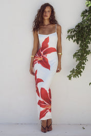 Abito lungo estivo bianco con stampa floreale rossa, spalline sottili, indossato da donna su sfondo bianco con pianta verde. Moda femminile elegante.