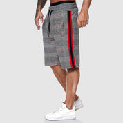 Shorts uomo a quadri grigi con strisce rosse e nere, elastico in vita, cordoncino nero, indossati con scarpe da ginnastica bianche.