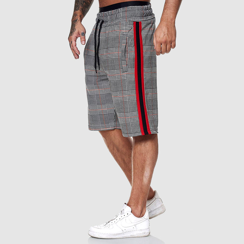 Shorts uomo a quadri grigi con strisce rosse e nere, elastico in vita, abbinati a scarpe da ginnastica bianche. Moda casual estiva.