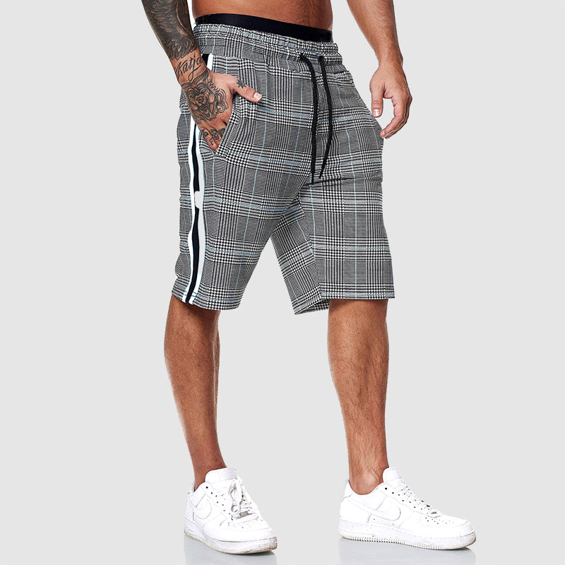Bermuda uomo a quadri grigi con strisce laterali bianche, elastico in vita e lacci, indossati con scarpe sportive bianche.
