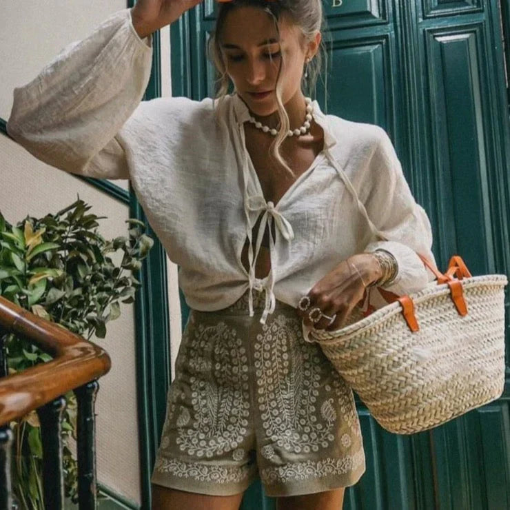 Donna con camicia bianca in lino, shorts ricamati beige, borsa di paglia intrecciata. Moda estiva, abbigliamento casual chic, accessori boho.