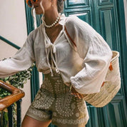 Donna con camicia bianca in lino, shorts verdi con ricami, borsa di paglia intrecciata, occhiali da sole arancioni, collana di perle, sfondo porta verde.