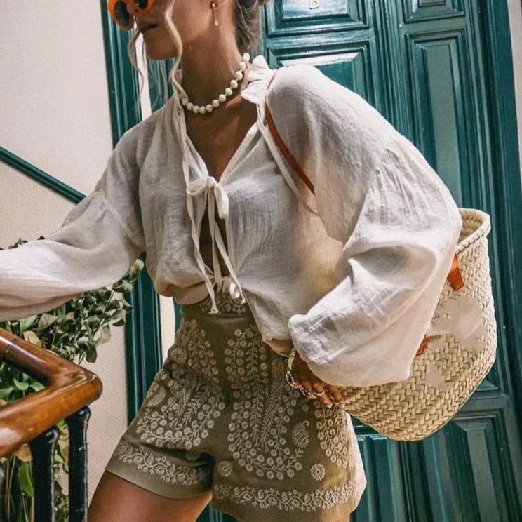 Donna con camicia bianca in lino, shorts verdi con ricami, borsa di paglia intrecciata, occhiali da sole arancioni, collana di perle, sfondo porta verde.