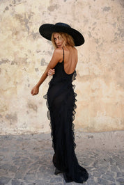 Donna in abito lungo nero trasparente con volant, cappello a tesa larga, posa elegante su sfondo murale. Moda estiva, abbigliamento femminile chic.