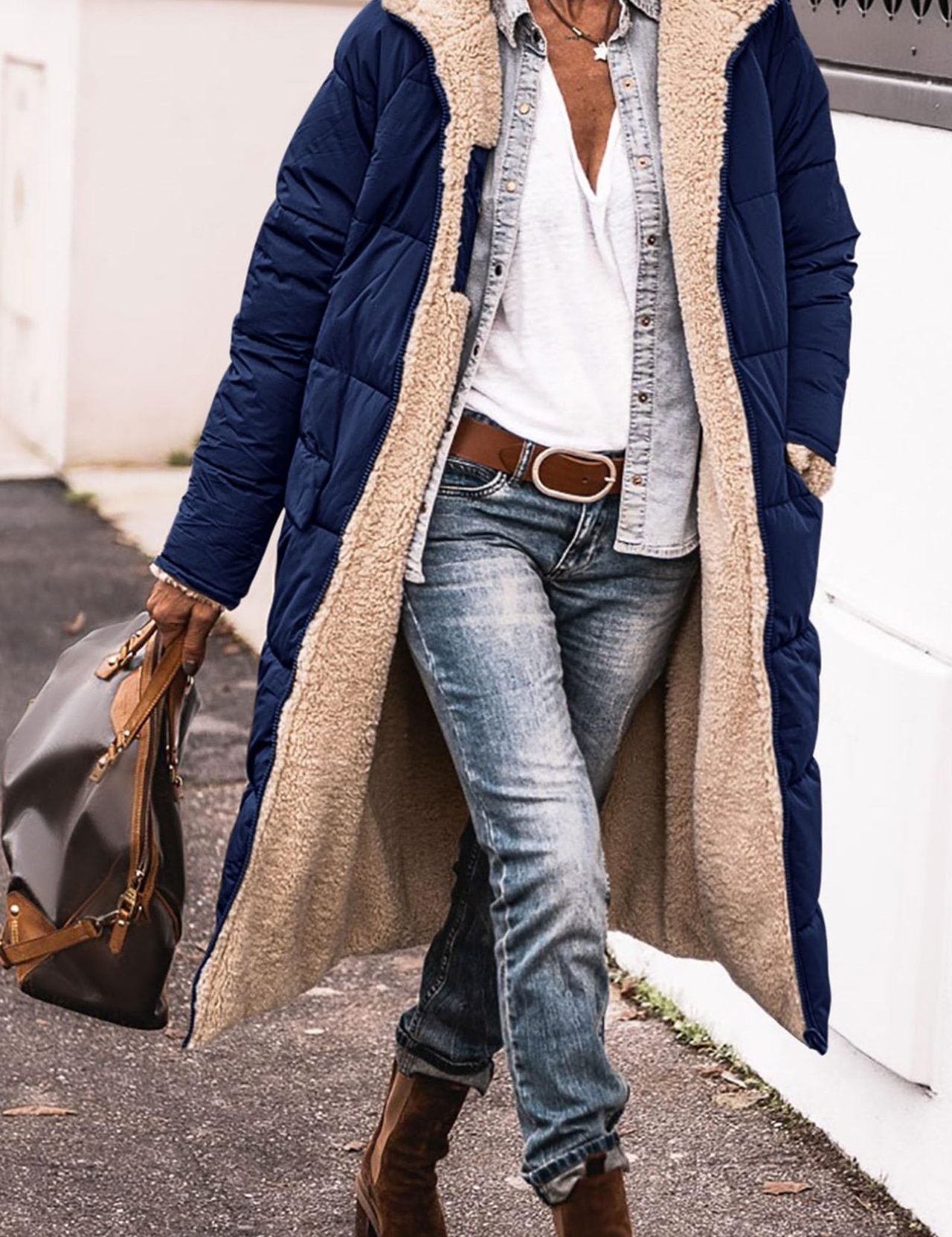 Donna con cappotto lungo blu imbottito, camicia bianca, jeans, stivali marroni e borsa in pelle. Moda invernale casual chic.