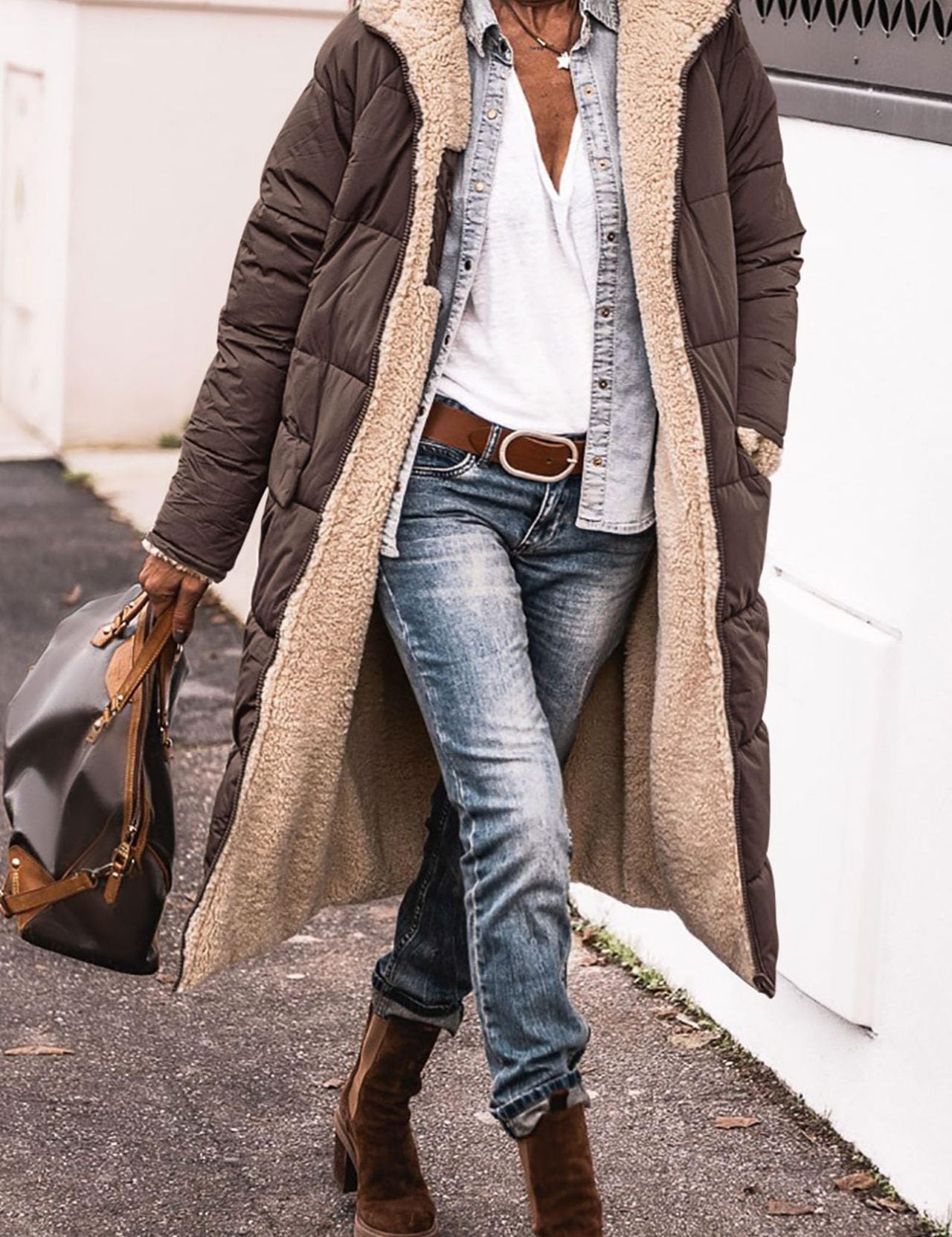 Donna con cappotto lungo marrone imbottito, jeans, stivali marroni e borsa in pelle. Moda invernale casual, abbigliamento caldo e confortevole.