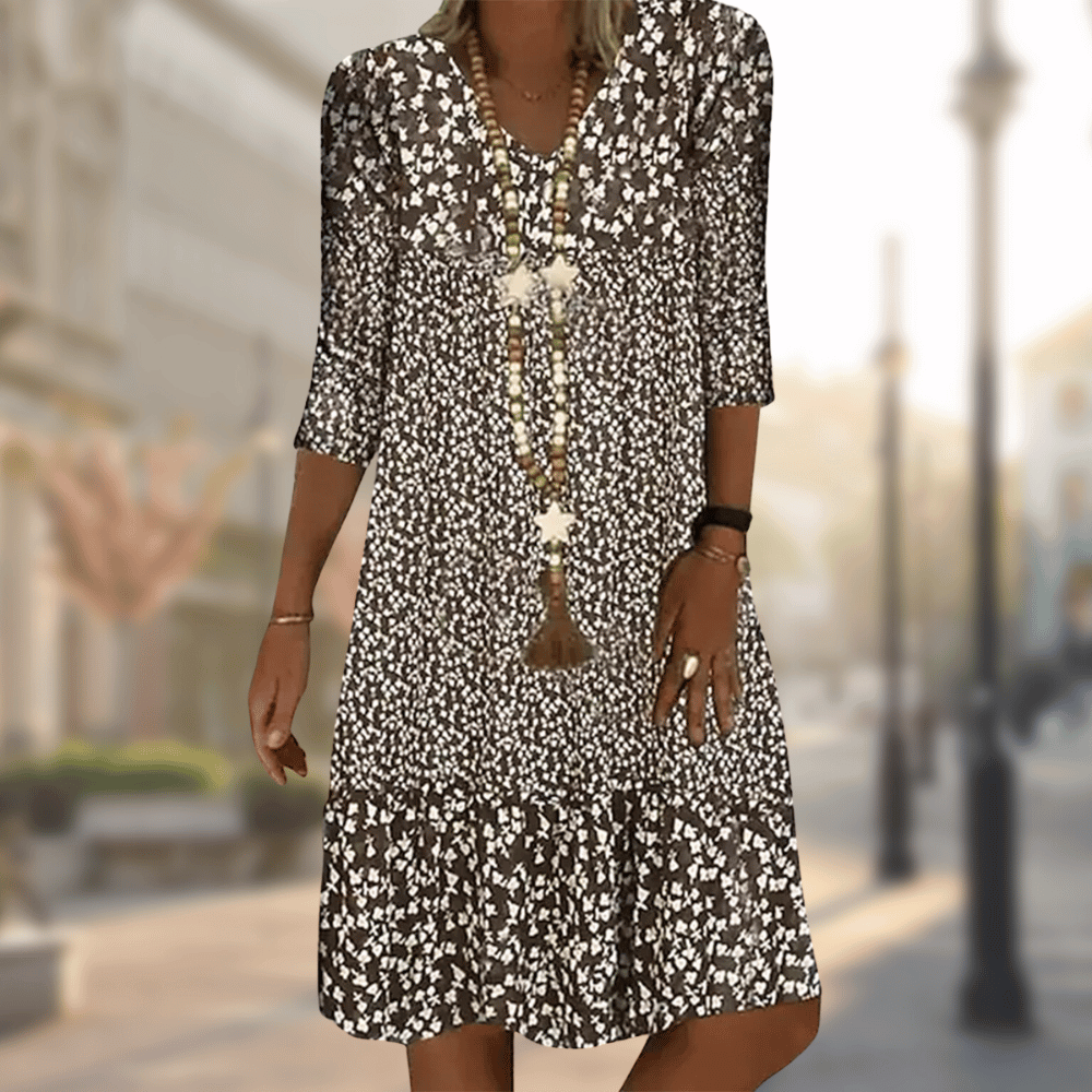 Abito estivo donna a fiori bianchi e neri, maniche a tre quarti, collana lunga, sfondo urbano sfocato. Moda casual elegante.