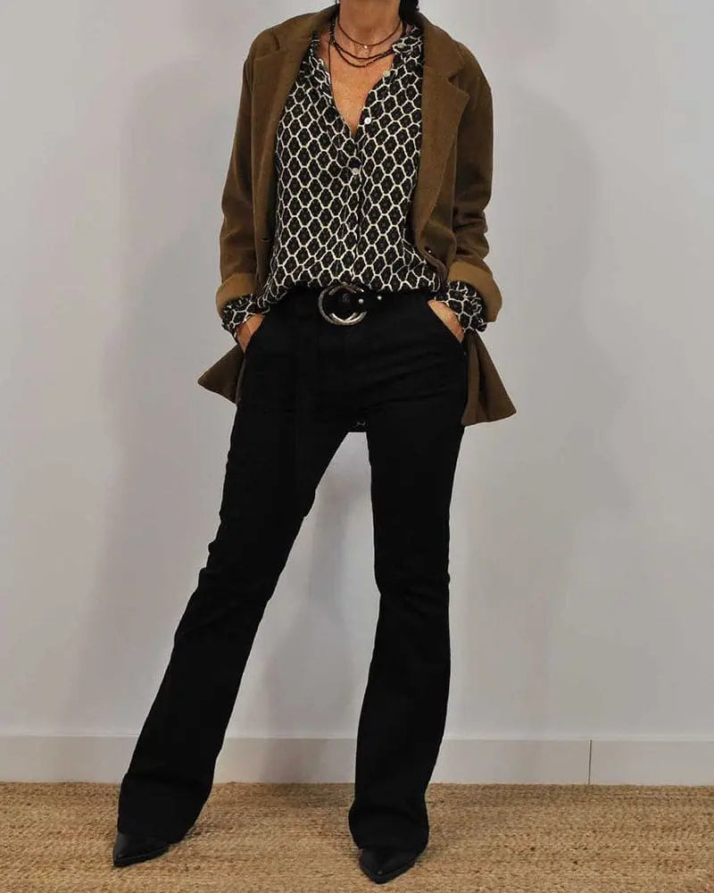 Giacca marrone su camicia a fantasia geometrica bianca e nera, pantaloni neri a zampa, cintura con fibbia grande. Look moda autunno elegante.