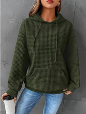 Donna con felpa verde a coste, cappuccio e tasca frontale, tiene una tazza da viaggio. Indossa jeans blu. Moda casual autunnale.