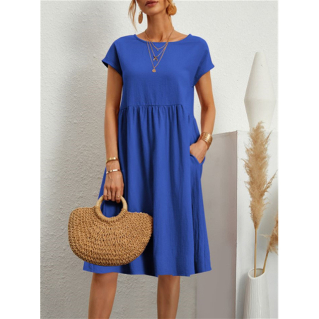 Abito estivo blu donna, maniche corte, con tasche. Accessori: borsa paglia, collane oro. Moda casual elegante. Sfondo bianco minimalista.