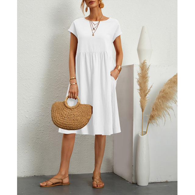 Donna con abito bianco estivo, sandali marroni, borsa di paglia, collane dorate. Sfondo minimalista con decorazioni in pampas. Moda casual elegante.