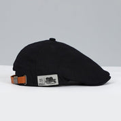 Cappello piatto nero in cotone con cinturino regolabile in pelle marrone, etichetta laterale, stile vintage, moda uomo, accessorio elegante.