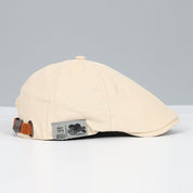 Cappello piatto beige in cotone con cinturino regolabile in pelle marrone, etichetta "Quality Fashion". Elegante accessorio moda uomo.