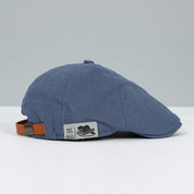Cappello piatto blu in cotone con cinturino regolabile in pelle marrone, etichetta "Genuine Fashion". Accessorio moda uomo elegante e casual.