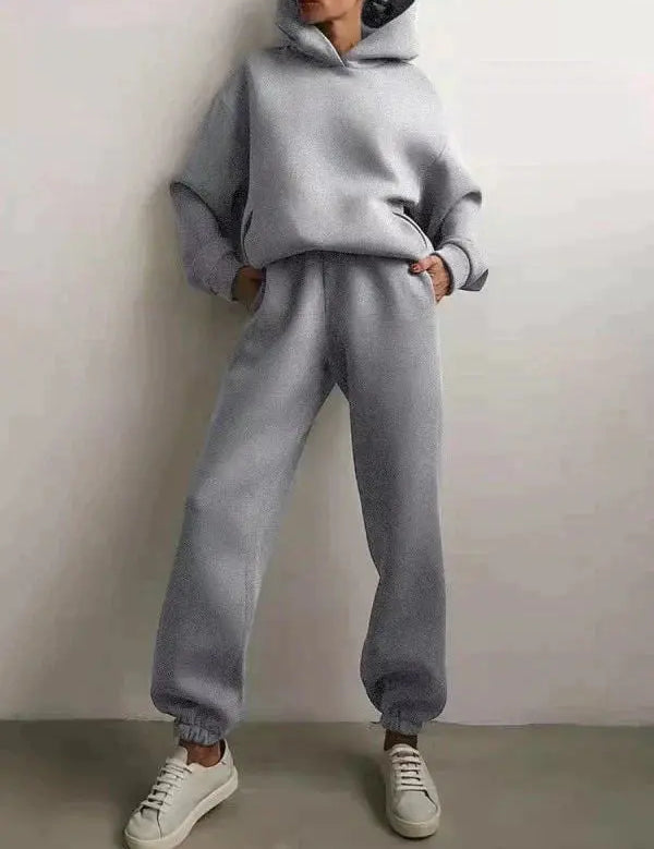 Donna in tuta sportiva grigia con cappuccio, pantaloni larghi e sneakers bianche. Abbigliamento casual, moda autunnale, stile confortevole.