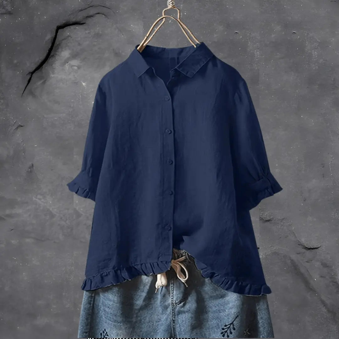 Camicia donna blu scuro in lino, maniche lunghe, colletto classico, abbottonatura frontale, orlo con volant, appesa su sfondo grigio.