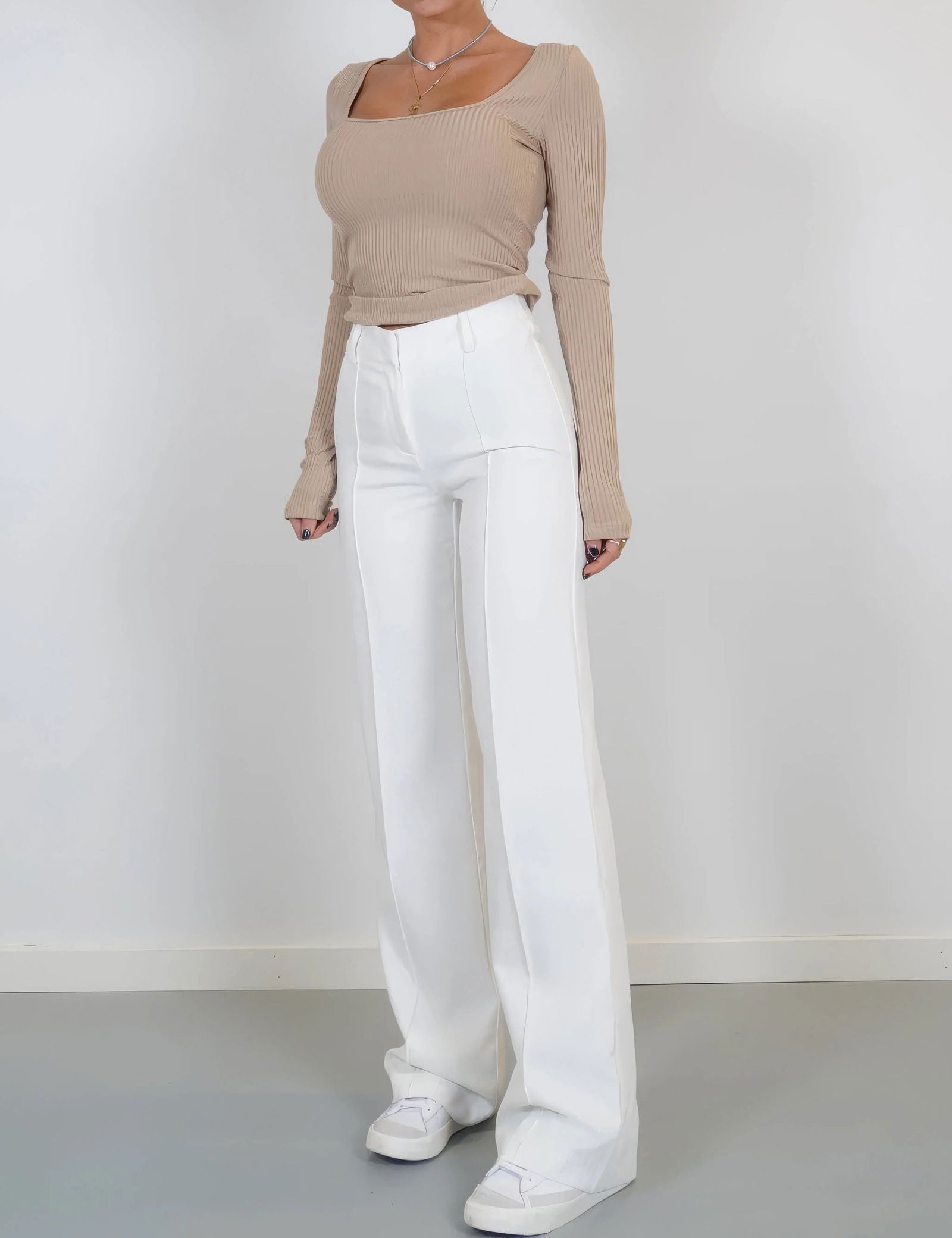 Donna con maglia beige a coste e pantaloni bianchi a vita alta, scarpe bianche. Moda femminile elegante, abbigliamento casual chic.