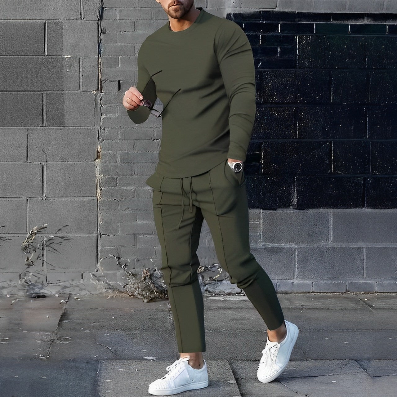 Uomo con completo sportivo verde oliva, maglia a maniche lunghe e pantaloni abbinati, indossa occhiali da sole e scarpe bianche, sfondo urbano.