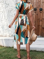 Donna con abito geometrico colorato, sandali marroni e cappello di paglia in mano, davanti a porta in legno. Moda estiva, abbigliamento casual.