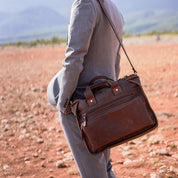 Uomo in abito grigio con borsa a tracolla in pelle marrone su sfondo naturale. Elegante borsa da lavoro, accessorio moda maschile.