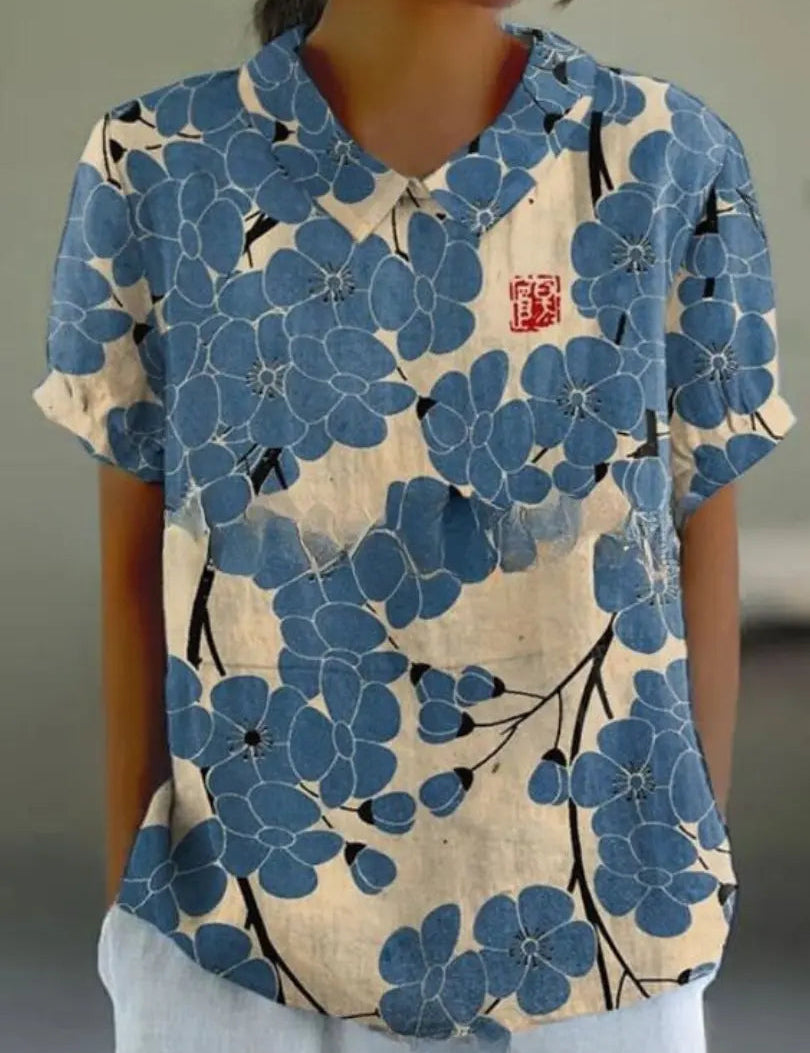 Camicia donna floreale blu, maniche corte, colletto classico, sfondo beige, design casual elegante, perfetta per moda estiva.