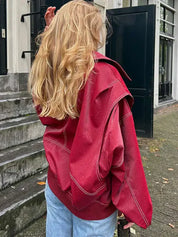 Giacca oversize rossa in pelle, indossata da una donna bionda di spalle, su sfondo urbano. Moda autunnale, stile casual chic.