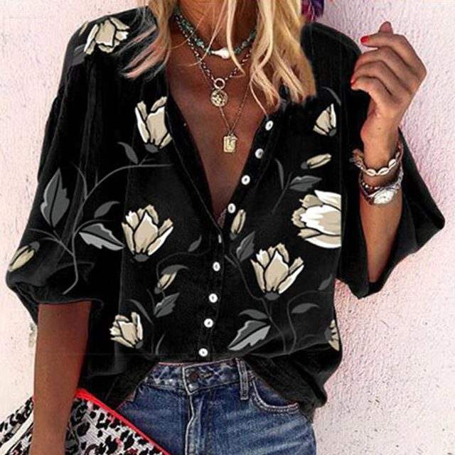 Camicia nera a fiori bianchi da donna, maniche lunghe, abbinata a jeans. Accessori: collane dorate e bracciale. Moda casual elegante.