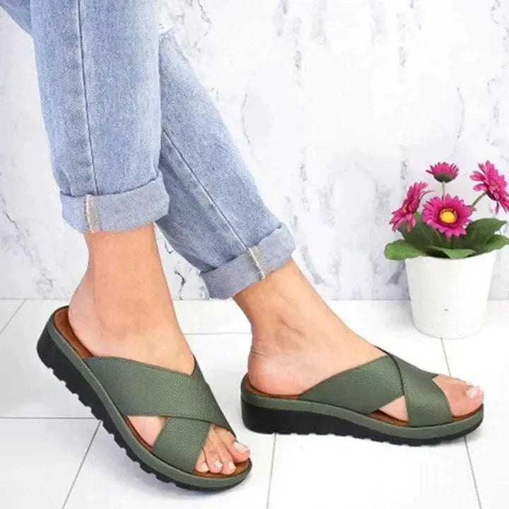 Jeans arrotolati e sandali verdi incrociati su suola nera, pavimento chiaro, vaso di fiori rosa. Scarpe estive comode, moda casual donna.