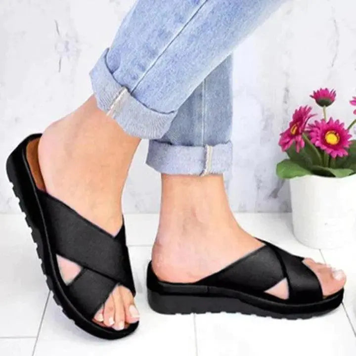Sandali neri incrociati da donna, suola antiscivolo, indossati con jeans chiari. Scarpe estive comode, moda casual, pavimento in marmo, fiori decorativi.