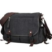 Borsa a tracolla in tela nera con chiusure a fibbia marroni, tasca frontale con zip, ideale per laptop e uso quotidiano. Stile casual e resistente.