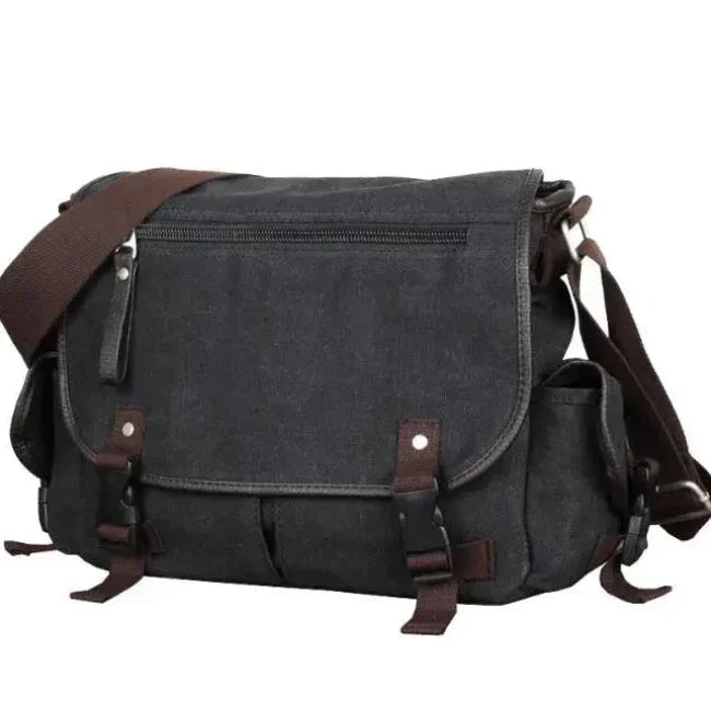 Borsa a tracolla in tela nera con chiusure a fibbia marroni, tasca frontale con zip, ideale per laptop e uso quotidiano. Stile casual e resistente.