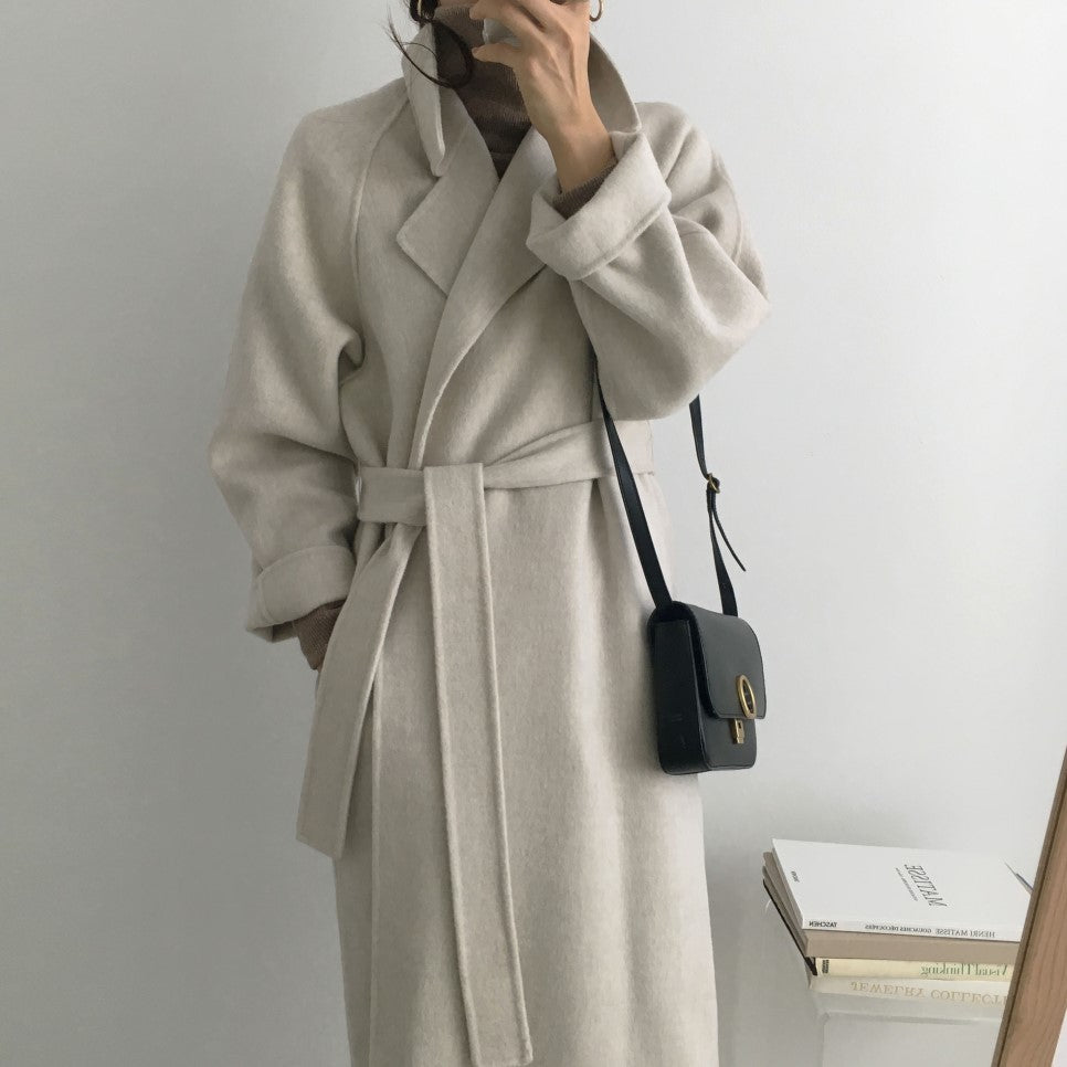 Cappotto lungo beige in lana con cintura, indossato da una persona. Borsa a tracolla nera. Moda invernale elegante e minimalista.