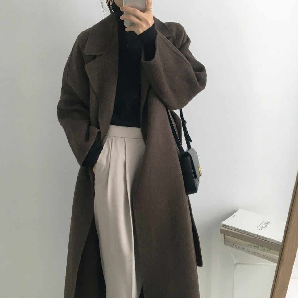Donna con cappotto marrone lungo, maglione nero e pantaloni chiari, borsa a tracolla nera. Moda autunnale elegante, stile minimalista.