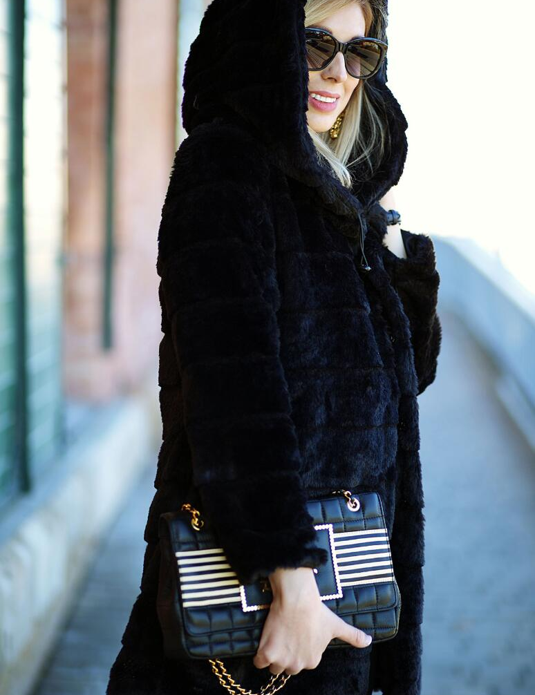 Donna con cappotto nero in pelliccia sintetica, occhiali da sole, borsa a tracolla nera con catena dorata e dettagli a righe. Moda invernale elegante.