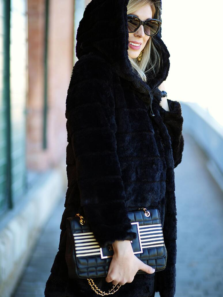 Donna con cappotto nero in pelliccia sintetica, occhiali da sole, borsa a tracolla nera con catena dorata e dettagli a righe. Moda invernale elegante.