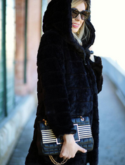 Donna con cappotto nero in pelliccia sintetica, occhiali da sole, borsa a tracolla nera con catena dorata e dettagli a righe. Moda invernale elegante.